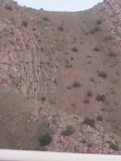 The image presents a high-angle or aerial view of a rugged, rocky landscape, consistent with a mountainous or hilly region like Tigranashen, Armenia. The terrain is characterized by earthy brown and grey rock formations, with sparse, dark green vegetation, likely bushes or small trees, scattered across the ground. In the mid-ground, subtle linear patterns, possibly ancient stone terracing or remnants of walls, are visible blending into the natural contours. The left edge of the image shows a bright, light-colored expanse, likely the sky, indicating a view from a significant elevation. The diffused lighting suggests it is daytime, possibly under overcast conditions, as shadows are not sharply defined. There are no discernible human figures or explicit activities taking place, and no visible text. The overall scene conveys a sense of a vast, natural, and potentially ancient environment. The image quality is somewhat blurry.