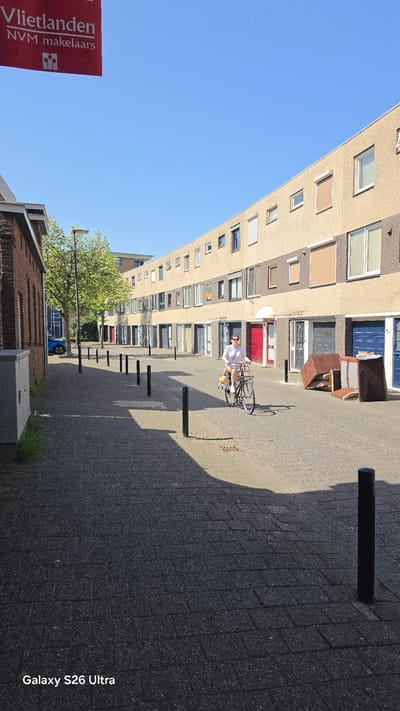 This is a landscape-oriented, vertical video of a residential street in Vlaardingen, Netherlands. The scene is dominated by a long, multi-story apartment building with light beige brickwork and numerous windows. In front of the building is a paved walkway with a series of dark bollards.
A person is riding a bicycle down the walkway, carrying a large, white, circular object on the back. The cyclist is wearing a light-colored shirt and shorts. The sun is casting distinct shadows, indicating it is likely daytime, with bright sunlight and clear blue skies, suggesting fair weather.
To the left of the frame, a red banner with white text reads "Vlietlanden NVM makelaars" and features a small logo. Further down the street, a leafy green tree provides a splash of color against the urban architecture. A blue car is parked in front of one of the buildings, and some red doors are visible on the apartment building.
The overall impression is a quiet, everyday scene in a European residential neighborhood on a sunny day. The video is oriented vertically, but the content is presented in a landscape format within the frame. The text "Galaxy S26 Ultra" is visible in the bottom right corner, likely a watermark from the recording device.