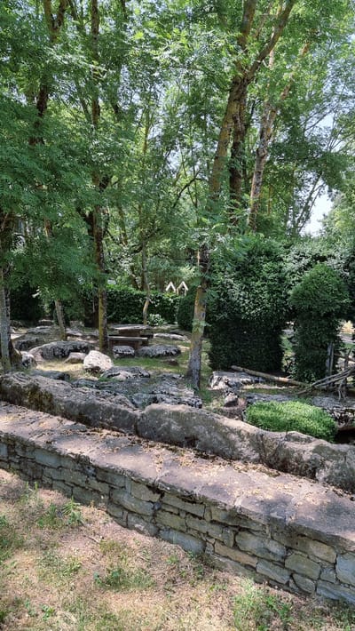 This media file depicts an outdoor scene with natural elements and a picnic table. The setting appears to be a park or garden area, characterized by lush green trees and a stone wall bordering a grassy area. The trees are tall and their branches are covered in leaves, suggesting a warm, sunny day, possibly late morning or early afternoon given the light.

The main subjects are the trees and the stone wall. The stone wall is constructed from irregular, weathered stones, suggesting it is old. In the middle ground, a rustic wooden picnic table with benches is visible, set amongst the rocks and trees. There are two decorative white birdhouse-like objects hanging from a tree branch, adding a whimsical touch to the scene.

The ground is covered in grass and scattered rocks. The overall atmosphere is serene and peaceful, evoking a sense of a relaxed outdoor setting. There is no visible text in the image. The location context suggests this scene is in Susqueda, Spain.