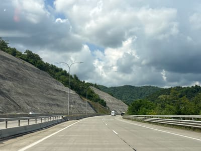 The image shows a highway with multiple lanes curving through a mountainous landscape. The road is paved with concrete, and white lines mark the lanes. On the left side of the road, a steep, rocky slope is reinforced with netting and stakes, with trees growing at the top. On the right side, the road is bordered by a metal guardrail and a concrete barrier. Lush green trees cover the hills in the distance, and a partly cloudy sky looms overhead. A white van and a dark vehicle are visible on the highway. The location is identified as a highway in Achara, Georgia.