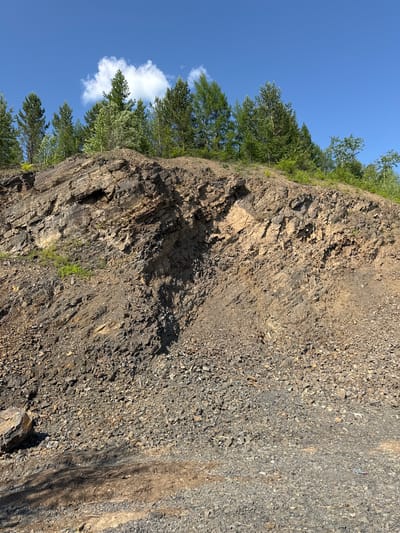 The image shows a steep, rocky cliff face under a clear blue sky. The cliff is composed of various shades of gray and brown rock, with some areas appearing more layered and fractured than others. A dense line of green trees and foliage runs along the top edge of the cliff, suggesting a natural landscape. A single white cloud is visible in the sky near the trees. The sunlight casts shadows on the cliff, indicating it is daytime. The texture of the rock ranges from large, jagged formations to smaller, gravel-like debris scattered across the surface. Some patches of green vegetation are also visible growing in crevices on the cliff face. The scene evokes a sense of rugged natural beauty.