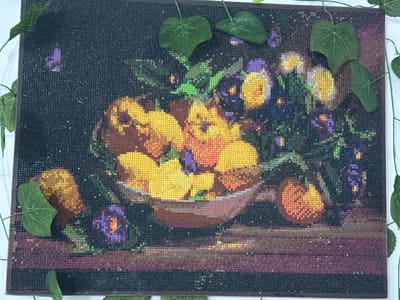 This is a diamond painting of a still life featuring a bowl of fruit and flowers. The artwork depicts a rustic wooden surface with a dark, textured background. In the center, a bowl overflows with yellow and orange fruit, possibly apples or peaches, interspersed with dark purple and vibrant yellow flowers that resemble pansies. The arrangement is further accented by additional scattered fruit and a single purple flower on the lower left. Green ivy leaves are strategically placed around the edges of the painting, adding a touch of naturalistic charm. The diamond painting technique creates a shimmering, textured effect due to the small, reflective beads. There is no indication of a specific location in Kojomkul, Kyrgyzstan, as the image is a piece of art and not a photograph of a real-world scene.