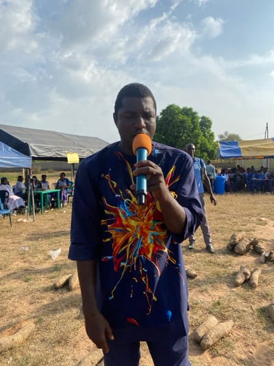 An outdoor scene in Itobe, Nigeria, depicts a man speaking into a microphone under a partially cloudy sky. The man, positioned centrally and in the foreground, wears a dark blue long-sleeved shirt with a vibrant, multicolored abstract splash design on the front. He holds a blue microphone with an orange foam cover to his mouth, with his eyes closed. His right hand has a ring on the fourth finger.
The ground is dry earth with sparse patches of grass. Scattered across the ground in the foreground and midground are multiple large, light-brown, irregular-shaped root vegetables, consistent with yams. A crumpled white plastic bag is visible on the ground to the left of the main subject.
In the background, to the left, a white and blue striped canopy tent is visible, with several individuals seated at tables underneath. Behind this, a darker, possibly black, tarp structure is erected. To the right background, another blue and yellow striped canopy tent is present, with more individuals seated in rows. A man in a light blue collared shirt and dark trousers stands facing right between the main subject and the tents. A large blue plastic barrel is also visible in the right background. Green foliage from trees is seen further in the distance against the horizon. The lighting suggests a bright, sunny day.