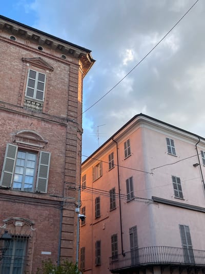 The image shows two buildings in the city of Piacenza, Italy, under a cloudy sky. The building on the left is made of red brick and features ornate architectural details, including a decorative cornice and a window with shutters. The building on the right is painted a pale pink and has a more modern appearance, with multiple windows also equipped with shutters and a balcony on the lower floor. A security camera is mounted on a pole between the two buildings. The scene is illuminated by the warm light of either sunrise or sunset, casting a gentle glow on the pink building.