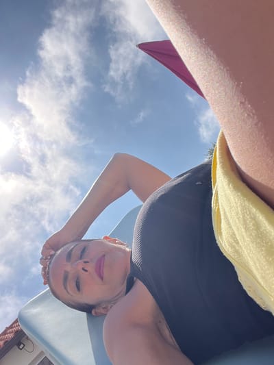 The image shows a woman lying on her back on a lounge chair. She is wearing a black top and a yellow towel is draped over her. The sky is blue with some clouds, and the sun is shining brightly. The woman's head is tilted back, and her eyes are closed. Her lips are painted with pink lipstick. Part of a purple umbrella is visible in the upper right corner of the image. The scene appears to be outdoors, possibly in a garden or by a pool. The overall impression is one of relaxation and enjoying the sunshine. There is no specific indication of the location within Caldas da Rainha, Portugal.