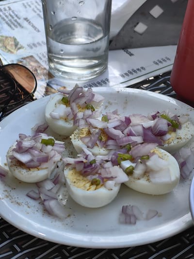 The image shows a white plate with several halves of hard-boiled eggs, each topped with chopped red onion and green chili peppers. The eggs appear to be seasoned with pepper. In the background, there is a glass of water, a menu, and part of a red bottle, possibly a ketchup bottle. The setting appears to be an outdoor cafe. There are no people visible in the image. The image quality is slightly grainy, and the lighting suggests it was taken during the daytime. The location is identified as Chhyal, India, based on context from the prompt, but there are no visual cues within the image itself to confirm this specific location.
