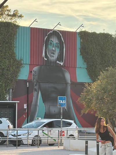 The image shows an outdoor urban scene in Platja D'en Bossa, Spain. A mural covers a corrugated metal wall, depicting a woman's face in shades of gray and turquoise, set against alternating stripes of red, turquoise, and burgundy. The mural extends down, depicting a figure in a dark jacket. Overgrown shrubbery partially obscures the base of the wall. A taxi stand is located adjacent to the mural. A blue "TAXI" sign is visible. Several white taxis are parked in the taxi stand. A young woman stands to the right of the taxi stand, wearing a black top and light-wash jeans.