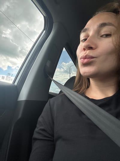 A medium close-up shot shows a woman inside a car with her seatbelt fastened. She is wearing a black t-shirt and is puckering her lips. The car interior is visible on the left and right sides, including a gray seatbelt stretching across her chest. Outside the car window, the sky is cloudy, with patches of blue visible. The scene is set in the city of Старое Русское Маматкозино, Russia.