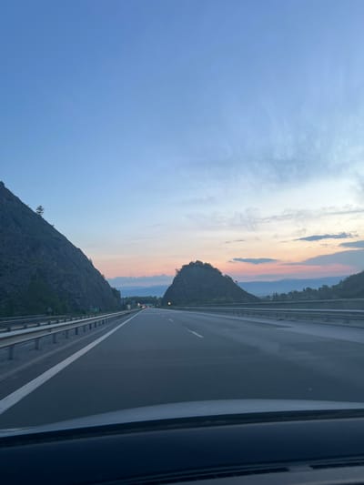The image depicts a road passing through a mountainous landscape at dusk. On the left, a steep, dark mountain rises sharply, sparsely covered with vegetation and a single tree. On the right, another, more rounded mountain is visible, also with sparse greenery. The road itself is a multi-lane highway with a clear white lane marker. Guardrails line the sides of the road. In the distance, the sky displays a gradient of colors from light blue at the top to pink and orange hues near the horizon, indicating sunset or sunrise. Clouds are scattered across the sky. The perspective suggests the photo was taken from inside a moving vehicle. Lights from distant traffic and possibly tunnels are visible on the highway ahead. The environment appears natural and undeveloped, with no visible buildings or significant man-made structures other than the highway infrastructure. The location is identified as the city of Malo Buchino, Bulgaria.