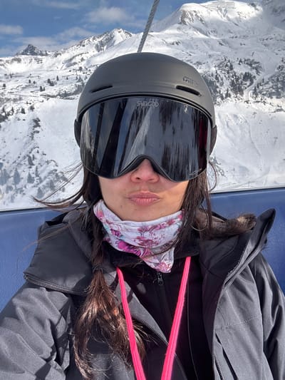 A close-up photograph depicts a single individual, identified as an adult female, seated within a ski lift in Zürs, Austria. The person wears a black Giro ski helmet and large, mirrored Sungod PKO Snow ski goggles, obscuring the upper face. A multi-colored neck gaiter with a floral pattern covers the lower face and neck, and the lips are pursed. The individual is dressed in a black ski jacket worn over a black zipped layer, with two distinct pink straps visible across the chest. A blue ski lift seat is partially visible behind the person. The background features a vast, sunlit, snow-covered mountainous landscape with multiple peaks, valleys, and sparse evergreen trees under a clear blue sky. A suspension cable, indicative of a ski lift, is visible at the top of the frame.