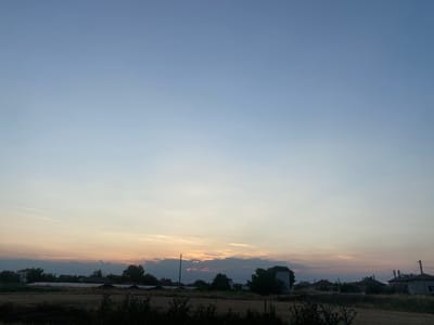 This is a landscape photograph of a rural setting in Kurtovo Konare, Bulgaria, likely taken at sunset or sunrise. The sky dominates the upper half of the frame, transitioning from a pale blue at the top to a soft, warm gradient of yellow and pink near the horizon, indicative of twilight. Wispy clouds are scattered across the sky, some catching the fading light.
The lower portion of the image depicts a village or small town. Silhouetted against the bright sky are the rooftops of houses, interspersed with trees. A large, white, greenhouse-like structure is visible in the mid-ground, suggesting agricultural activity. In the foreground, dry, golden grass and dark foliage are visible, hinting at a field or undeveloped land. A single utility pole stands prominently in the middle of the frame, with wires stretching across the scene.
The overall mood is serene and peaceful, characteristic of a quiet evening or early morning in the countryside. The lighting suggests a time of day when natural light is soft and diffused. There is no visible text in the image.