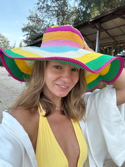 A woman is posing for a selfie on a beach. She is wearing a yellow bikini top and a white shirt draped over her shoulders. She has on a large, colorful crocheted sun hat with stripes of pink, orange, yellow, light blue, white, and green. The woman has blonde hair and is smiling at the camera. In the background, there is a sandy beach, trees, and a rustic wooden structure with a corrugated metal roof. The scene appears to be outdoors, possibly in a tropical location. There is no indication that this location is in Thuận Thành, Vietnam.