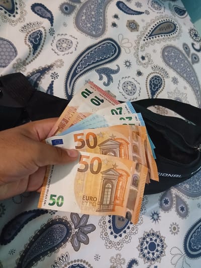 This image shows a hand holding several euro banknotes of different denominations on top of a black bag on a paisley-patterned surface. The banknotes include a 10 euro note, a 20 euro note, and several 50 euro notes. The bag is partially visible, with a strap and zipper. The surface appears to be a bed or possibly a fabric surface with a repetitive paisley design in blue, grey, and white. The image does not contain sufficient information to determine the location as Vetren Dol, Bulgaria, or any other geographical location. There are no people present in the image except for the hand.