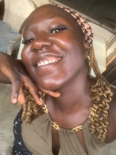 A close-up, high-angle selfie shows a Black woman with dark skin, a patterned headscarf, and braided hair smiling. She is wearing a tan top with a gold chain detail around the neckline. Her left hand is resting on her chin, and her right arm is bent, with her hand near her face. The background appears to be outdoors, with a concrete floor and some indistinct structures. The image is slightly blurry due to the close proximity and motion of the camera. It is not possible to determine the specific location in Eliozu, Nigeria based on the visual information alone.