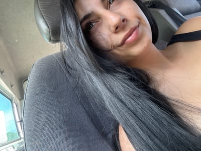 A woman with long, dark hair is pictured inside a vehicle. She is sitting in a gray upholstered seat, with the headrest visible behind her. Her complexion is light and she has dark eyes. She is wearing a black top. Part of a window can be seen in the background, along with a slightly visible environment suggesting an outdoor setting. There is no indication of Strumyani, Bulgaria, or any other specific location.