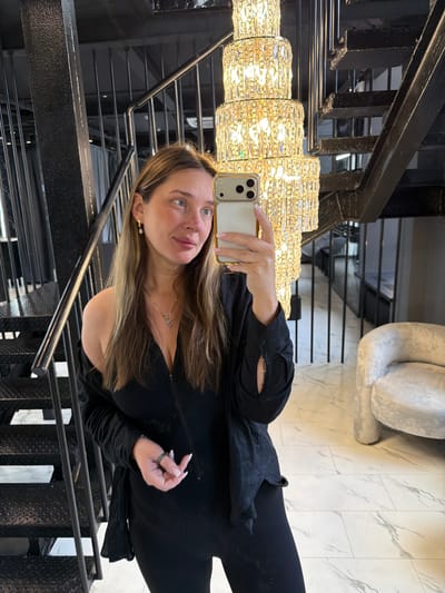A woman is taking a mirror selfie in what appears to be a modern staircase and living area. She is standing near a black metal staircase with open risers, and a large, multi-tiered crystal chandelier hangs above and to her right. The floor is covered in large, marble-patterned tiles. To her right, a light gray, textured armchair is visible. The woman is wearing a black, off-the-shoulder top with long sleeves and a silver necklace with a cross pendant. She is also wearing a green ring on her right hand. Her hair is long and brown, and she is holding a smartphone in her right hand, angled to capture the reflection. The location is in the city of Укромновское сельское поселение, Ukraine.