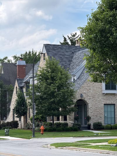 The image shows a large, modern brick house with a dark gray roof, viewed from the street. The house has several windows and an arched entryway with a dark door. Trees and manicured green lawns surround the property, with a sidewalk and street visible in the foreground. An orange fire hydrant stands on the lawn near the street. The sky is partly cloudy, suggesting daytime weather. The scene is a residential street in Naperville, United States of America. There is no visible text in the image.