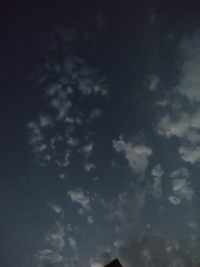 The image depicts a dark, cloudy sky, likely taken at dusk or night. There are scattered white clouds against a deep blue to near-black sky. A small, faint point of light, possibly a star or distant airplane, is visible in the upper left quadrant. The lower portion of the image shows the silhouette of a dark structure, possibly a rooftop or the top of a building, with a triangular peak. The scene is devoid of any visible people or specific landmarks that would pinpoint a precise location within Арчалы, Kyrgyzstan. The overall impression is one of a quiet, possibly overcast evening sky over a sparsely populated area.