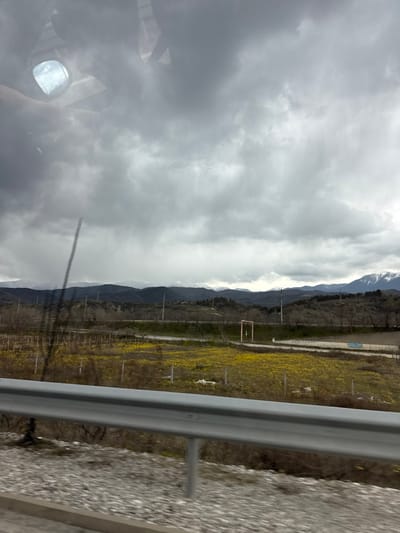 The image depicts an expansive rural landscape in Strumyani, Bulgaria, viewed from a moving vehicle, evidenced by motion blur in the foreground and reflections on the upper left glass surface. The sky is predominantly overcast with dense, grey cloud formations. In the immediate foreground, a silver metal guardrail runs horizontally across the lower third, positioned above a gravel or stone road shoulder.
Beyond the guardrail, a vast field dominates the midground, characterized by a vibrant spread of yellow flowering vegetation. Scattered within this field are numerous thin, dark posts, possibly remnants of fencing or agricultural stakes. A dirt or unpaved track traverses the midground horizontally, separating the yellow field from further vegetation. Along this track and slightly above it, multiple utility poles with overhead wires are visible. To the right of the frame, past the track, a reddish-brown, rectangular frame structure stands upright, and further right, a light-colored embankment with indistinct blue graffiti is present.
The background features a prominent range of dark, undulating mountains, some of which display distinct snow-capped peaks, particularly towards the right side of the frame. The overall impression is a cloudy day in a mountainous agricultural region. No individuals or specific actions are discernible.
