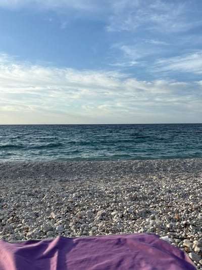 The image displays a coastal scene, likely taken at a beach in Portoferraio, Italy, divided horizontally into three distinct sections: the sky, the sea, and the pebble beach.

The upper third of the frame shows a light blue sky adorned with scattered, wispy white clouds, suggesting a partly cloudy day. These clouds appear to be high-level cirrus or altocumulus, creating delicate patterns across the expanse of blue.

The middle section features the ocean, characterized by its deep blue-green hue. The surface of the water is textured with numerous small waves, indicating gentle movement and creating a shimmering effect. The horizon line separating the sea and sky is visible but indistinct due to the soft lighting and distant haze.

The lower third of the image captures a pebble beach. The beach is densely covered with countless small, rounded pebbles, predominantly in shades of light grey and white. The lighting, possibly from late morning or afternoon sun, casts subtle shadows among the stones, enhancing their three-dimensional texture. On the far right side of the frame, a vibrant purple fabric, likely part of a towel or clothing, is visible, partially covering the pebbles and hinting at human presence, though no people are shown.

The scene conveys a calm and peaceful atmosphere. There is no specific activity taking place, rather a tranquil snapshot of the natural environment. No visible text is present in the image.