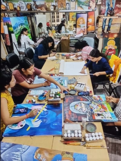 The image depicts an active art studio or classroom located in Aktash, Uzbekistan. Approximately nine individuals, predominantly young adults, are seated at long, light-colored tables, actively engaged in creating art.
In the foreground, from left to right: a person in a yellow patterned top with glasses is painting a large blue canvas featuring abstract white hands and faces using a yellow-handled brush; a person in a maroon t-shirt is reaching towards a blue canvas; a person in a dark blue top is drawing or sketching on a large white paper roll; and a person in a striped top (partially visible) is painting a canvas with a colorful, abstract portrait. The tables are covered with various art supplies, including numerous paint pots, palettes, brushes, pencils, rulers, and water containers. Several canvases are in different stages of completion.
The midground and background reveal more participants seated at additional tables, also focused on their artworks. The walls are densely covered with a wide array of completed or in-progress paintings, showcasing diverse styles and subjects such as landscapes, portraits, and abstract art. Shelves above the workstations display more art, trophies, and decorative objects. A multi-paned window is visible in the background, illuminating the indoor artistic workspace.