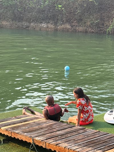 A woman and a man are seen on a wooden pier extending into a body of water. The woman, wearing a red floral dress, is reaching towards the man. The man is sitting on the edge of the pier, wearing a red life jacket, with his feet dangling in the water. A blue volleyball is floating in the water near them. The pier is constructed of wooden planks. The water appears greenish. Vegetation is visible on the shoreline opposite the pier. The scene is within Tambon Nam Phrae, Thailand.