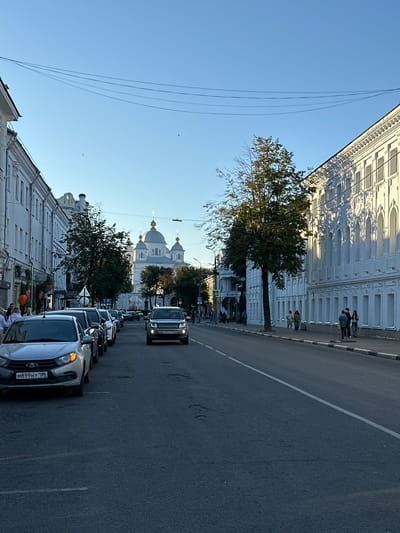 The image depicts a street scene in Yazvitsevo, Russia, under a clear blue sky. On the left side of the street, several white buildings with decorative facades line the road. Some of these buildings have multiple windows and architectural embellishments. A row of cars is parked along the side of the street, with the closest vehicle being a silver sedan. Further down the street, a white SUV is in motion.
On the right side of the street, a long, white building with a highly ornate facade, featuring numerous arched windows, dominates the view. A large tree with green and reddish leaves stands between the building and the road. Several people are visible walking on the sidewalk in front of this building.
In the distance, at the end of the street, a large white church with multiple golden domes is prominently featured. The street itself is asphalt with white lane markings. Power lines are visible in the sky, strung between poles. The overall impression is a daytime street view in a Russian town or city.