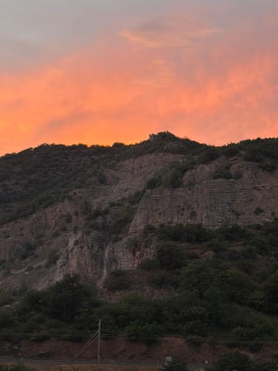 This image captures a dramatic mountain landscape at sunset. The sky is filled with vibrant orange and pink clouds, indicating the golden hour. On the right side of the frame, a steep, rocky mountain face rises, covered in dense green vegetation. The texture of the rock is rough and layered, suggesting ancient geological formations. Along the base of the mountain, a railway line is visible, marked by tracks and utility poles, hinting at human infrastructure within the natural environment. The lighting suggests either sunrise or sunset. The overall mood is one of natural beauty and grandeur, with a touch of human presence. There is no visible text in the image.