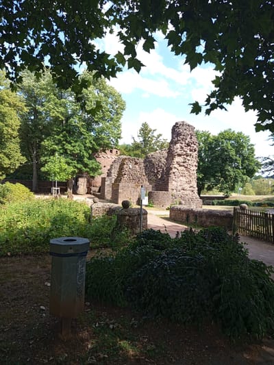 A sunny day is captured in this image, with the sky showing a mix of blue and white clouds. The scene features the ruins of an old stone structure, possibly a castle or a fortification, with crumbling walls and towers made of rough-hewn stone. Lush green foliage surrounds the ruins, with trees in the background and dense bushes in the foreground. A weathered, octagonal trash can with some graffiti is visible in the lower left of the image. To the right, a rustic wooden fence lines a path. The overall setting appears to be a park or historical site in Illingen, Germany. No people are present in the frame. The time of day appears to be daytime, given the bright lighting and clear sky.