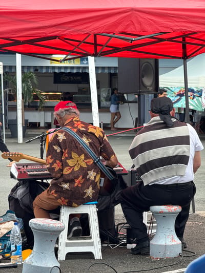 The image captures an outdoor musical performance or rehearsal in Noumea, France. Two men are seated, facing away from the camera, under a large red canopy that occupies the upper portion of the frame. The man on the left, wearing a brown short-sleeved shirt with yellow, orange, and white floral patterns, a red baseball cap, and brown trousers, sits on a white plastic folding stool. He is playing an electric guitar, secured by a blue and white patterned strap, and has a red and black electronic keyboard positioned on a stand in front of him. A microphone stand is visible near the keyboard. To his right, a second man is seated on a grey, weathered bollard-like stool with orange paint remnants. He wears a white collared shirt, black trousers, and a black head covering tied at the back. A grey and black striped sweatshirt is draped over his shoulders.
Between the performers and the background, a large black audio speaker is mounted on a stand. In the mid-ground, a blue line, possibly a rope or barrier, extends horizontally. The background features a storefront with the sign "Le Sésame" and an "ENTRÉE" sign with a downward arrow visible above its entrance. Display cases with food items are visible inside the shop, where a woman in a light denim jacket and white skirt is standing. Palm tree foliage is visible near the storefront to the left. The ground surface is paved, and black cables are visible, along with a blue plastic water bottle near the left performer's feet. Another individual is partially visible behind the speaker in the right background.