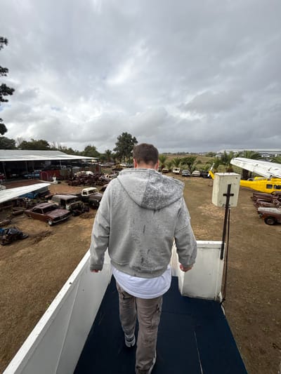 The image shows a person standing on a white and blue surface, possibly a structure or platform, looking out over a scene with numerous old cars and vehicles, and a yellow helicopter. The location is Kraaifontein, South Africa. Overcast sky.