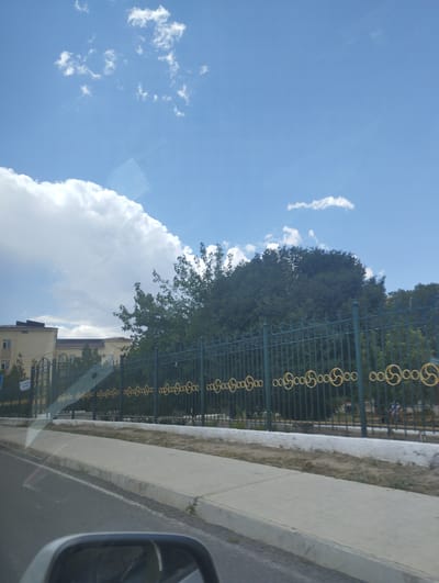An eye-level, medium shot captures a daytime scene from inside a vehicle in Forish QFY, Uzbekistan. The foreground is dominated by the reflection in the car's side mirror, showing a portion of the vehicle's interior and the asphalt road. A concrete curb and sidewalk are visible to the right of the road. Beyond the sidewalk, a dark green metal fence with a decorative gold pattern runs horizontally across the frame, separating the viewer from the background. Behind the fence, a line of dense green trees creates a natural barrier. Further in the background, a light-colored, multi-story building with a dark roof is visible. The sky above is a bright blue, dotted with scattered white clouds, some of which are large and fluffy, suggesting fair weather. The overall impression is one of a typical urban or suburban streetscape under a clear sky.