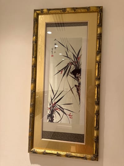 A framed artwork depicting orchids and Chinese calligraphy hangs on a beige wall. The frame is golden with a bamboo-like texture. The artwork is a vertical composition with dark green orchid leaves and small red flowers against a light background. There are two white circular reflections on the glass of the frame. The wall appears to be a textured plaster. The overall scene is indoors, likely in a residential or commercial setting. No specific location in Сосны, Russia can be determined from the image.