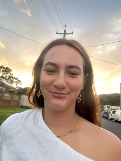 A young adult female is centered in a mid-shot image, facing forward with a faint, closed-mouth smile. She wears a white off-the-shoulder garment, gold-colored snake-shaped dangle earrings, and a delicate gold-colored necklace featuring small green beads. Her long brown hair is visible, illuminated by warm, low-angle sunlight from the left rear, suggesting either dawn or dusk.

The background depicts a suburban street scene located in Thirroul, Australia. To the subject's left, a residential house with solar panels on its roof and a low brick wall is visible behind greenery. To the right, a paved street extends into the distance, lined with several parked vehicles, primarily white vans or utility vehicles. Distant hills and a treeline are discernible beyond the street. A utility pole with multiple overhead power lines diagonally traverses the upper portion of the frame against a partly cloudy sky, exhibiting warm orange and yellow hues near the horizon transitioning to lighter blue.