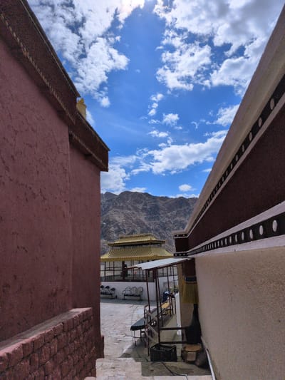 This is a wide shot of a courtyard in Choma, India, with mountains in the background. The sky is bright blue with fluffy white clouds. Two walls, one red and one cream-colored, frame the shot, creating a path that leads to a golden-roofed building in the distance. Several benches are visible in the courtyard, and a person can be seen sitting on one of them. A black crate is on the ground next to a bench. The image appears to have been taken on a sunny day.