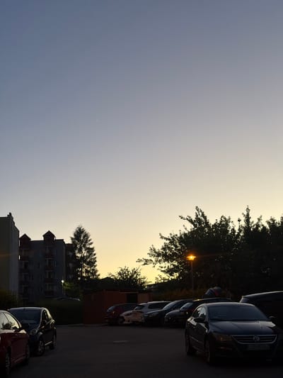 The image depicts a parking lot in Lublin, Poland, during twilight. In the foreground, several cars are parked. A dark-colored sedan with license plate "LUBL 90162" is prominently positioned on the right. To its left, a dark hatchback is visible, and further to the left, the rear of a red car. In the background, a multi-story apartment building with distinctive architectural features, including peaked roofs on several sections, stands. Adjacent to the building is a tall evergreen tree. Beyond the cars and building, a line of dense trees is silhouetted against the darkening sky, with a streetlamp illuminated among them. The sky transitions from a pale blue at the top to a warm yellow hue near the horizon, indicating the sun has recently set. There are no people visible in the scene.