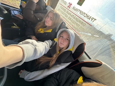 Two young women are standing inside a vehicle, possibly a bus, as suggested by the window and seating visible. The women are wearing hooded sweatshirts and jackets. The text on the exterior window reads "MYSI", indicating the location is likely Mysy, Russia. The environment appears to be outdoors, based on the outside reflections and signs on the glass. The women are positioned close to the camera, presumably taking a selfie.