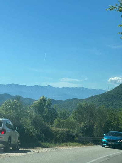 A bright, clear sky dominates the upper portion of the image, with a faint contrail from an aircraft visible. Below the sky, a vast mountain range stretches across the horizon, its peaks layered and receding into a hazy blue. The mid-ground is a tapestry of lush, green hillsides covered in dense vegetation. In the foreground, a paved road curves to the right, with a gravel shoulder. A silver SUV is parked on the shoulder, partially obscured by trees on the left side of the frame. On the right side of the road, a bright blue sedan is driving away from the camera, its license plate visible. Tall, green trees and bushes line the side of the road, with some flowering plants interspersed. In the background on the right, several tall power line towers are visible against the green hillside and sky. The scene appears to be a scenic overlook or a mountain road.