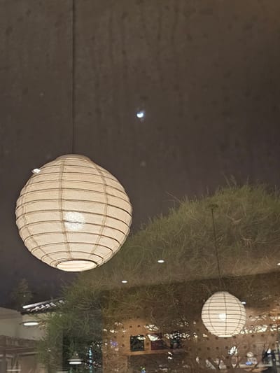 The image depicts an indoor scene at night, possibly a restaurant or cafe, reflected in a window. The primary subject is a large, round, white paper lantern hanging from the ceiling, illuminated from within. To the right and behind it, another smaller paper lantern is visible. The background appears to be a dark ceiling with a faint reflection of what might be a crescent moon or a light source. A dense, green shrubbery is reflected in the window, creating a blurred backdrop. The reflection also shows the interior of the establishment, with hints of architectural details and scattered lights. The scene is static and appears to capture a moment of quiet ambiance. The image does not provide any specific information about the city of Ngaglik, Indonesia, beyond the possibility of it being the setting.