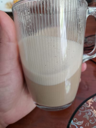 A clear glass mug, partially filled with a layered beige and brown liquid, is held in a person's hand. The mug has vertical ridges along the exterior. The liquid appears to be a type of coffee drink, possibly a latte or cappuccino, with layers of milk or foam. The background is a brown surface, partially obscured by the hand and the mug. A decorative plate with a pattern in silver tones is visible in the upper right corner.
There is no information to determine the location is Bochum, Germany.