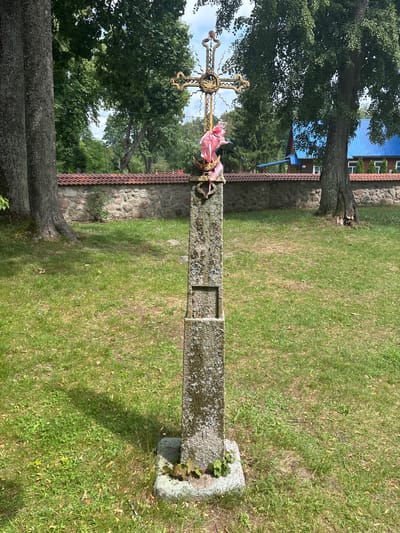 The image depicts a weathered stone monument with a decorative metal cross at its apex, situated in an outdoor setting that appears to be a cemetery or churchyard in Puchły, Poland. The stone base of the monument is rough-hewn and moss-covered, with a visible square indentation on its front. A bright pink ribbon is tied around the cross, suggesting a recent adornment.
Behind the monument, a low stone wall with a terracotta tile roof runs horizontally across the frame. Beyond the wall, a building with a bright blue roof is visible, likely a church or another structure associated with the location. Large trees with lush green foliage frame the scene, with their trunks on the left and right sides of the image. The ground is covered in green grass, interspersed with fallen leaves and small wildflowers. The sky above is partly cloudy, with patches of blue peeking through. The overall impression is one of a tranquil, perhaps historical, rural site.