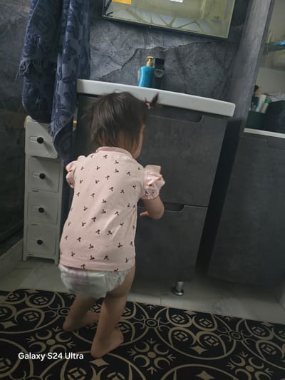 The image shows a toddler from the back, wearing a pink shirt with a pattern of small brown bows and a diaper. The child is standing on a patterned rug with black and gold designs. The setting appears to be a bathroom, with a sink visible to the left and dark cabinets behind the child. A blue bottle with a yellow pump dispenser is also present on the counter. The lighting suggests it is daytime. On the right side of the image, text is visible: "Galaxy S24 Ultra". The context provided indicates the location is Tilekmat, Kyrgyzstan.