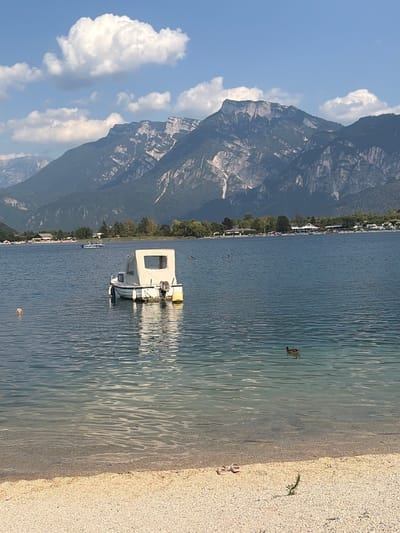 This is a vertical, high-angle, eye-level shot of a tranquil lake with a sandy shore. A white boat with a canvas cover is moored in the clear blue water, its reflection rippling on the surface. The boat has a small window and a yellow buoy attached. In the distance, a line of trees and buildings, possibly a village, runs along the shoreline. Majestic mountains with rocky faces and patches of green vegetation dominate the background. The sky is a clear blue with a few fluffy white clouds. On the sandy beach, a pair of pink sandals are visible, along with a small sprig of green vegetation. The scene suggests a peaceful, sunny day in a lakeside resort, likely in the late afternoon given the soft light and shadows. The water is calm, with gentle waves lapping the shore. A duck can be seen swimming in the water to the right of the boat. Another, smaller boat is further in the distance, closer to the shore. The overall mood is serene and inviting, evoking a sense of relaxation and natural beauty.