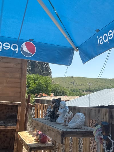 An outdoor scene in Bjni, Armenia, is depicted. The foreground is dominated by a large blue Pepsi umbrella, casting shade over a wooden structure. On a shelf of this structure, several small statues are displayed, including an angel with its hands clasped, a smaller statue of a figure, and what appears to be a pair of angel wings. To the left of the statues, a red object wrapped in plastic is visible on a lower shelf. In the background, a rugged, rocky cliff face rises behind lush green trees. Further in the distance, rolling green hills form the horizon under a clear blue sky. The overall impression is of a casual, outdoor setting, potentially a market or roadside stall, with a backdrop of natural Armenian landscape.