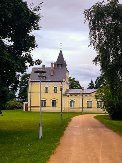 The image depicts a view of the Salaspils Manor House (Salaspils muiža) located in Salaspils, Latvia. The manor house, painted a pale yellow, features a dark pitched roof with a prominent tower topped with a weather vane. Several windows with arched tops are visible on the facade, along with decorative brickwork above some of them. The building is flanked by lush green trees, with the leaves of a large deciduous tree framing the left side of the shot and a tall birch tree dominating the right. A gravel pathway curves away from the viewer towards the manor, bordered by a neatly mowed green lawn. Two modern lampposts with disc-shaped tops are positioned along the pathway, and another lamppost is visible in the foreground on the lawn. The sky is overcast with scattered clouds. There are no people or animals visible in the scene.