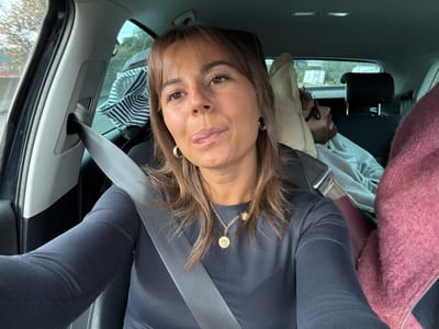 The scene is set inside a car in Aix-en-Provence, France. A woman occupies the front passenger seat, secured by a grey seatbelt. She has brown hair and is wearing a dark long-sleeved top, gold hoop earrings, and a gold necklace with two circular pendants. Her expression features her tongue extended towards the camera. In the rear passenger seat, partially visible behind the woman, a man with a beard and sunglasses is observed resting or sleeping, with his head supported by a beige cushion or headrest. He is dressed in a light grey hoodie. A striped garment is visible in the backseat behind the woman, and a fuzzy, reddish-brown object, possibly another garment, is on the right side of the rear seat. The exterior environment, visible through the car windows, consists of blurred trees and foliage, indicating the vehicle is in motion during daylight hours.