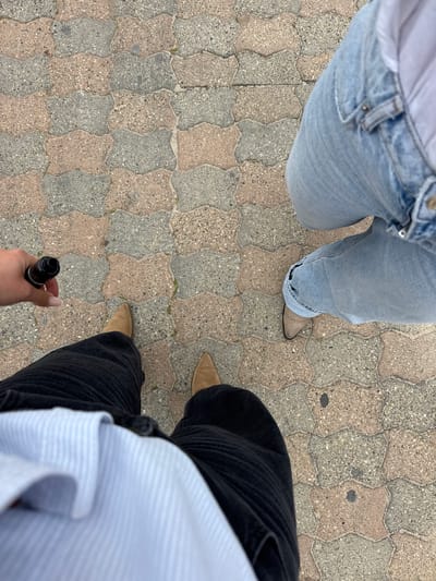 The image shows a view from above, looking down at two individuals standing on a paved surface comprised of interlocking, geometrically shaped stones in a light gray and tan color. The person on the left is wearing light-wash blue jeans and tan-colored pointed-toe boots. The person on the right is wearing black pants and tan pointed toe boots, and is holding a black cylindrical object in their right hand. The person on the right is wearing a light blue and white striped shirt, or partial shirt. The location is unspecified, and there is no way to determine if it is in the city of Vallauris, France. The pavement is a mix of two colors and the lighting of the scene is natural. There is no indication of any specific action or interaction.