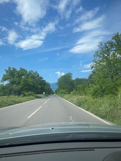 The image shows a perspective from inside a car driving on a rural road. The road is paved, with a white line dividing lanes. On either side of the road are grassy verges and lush green trees. The sky is bright blue with scattered white clouds. In the distance, a mountain range is visible. The hood of the car in the foreground features a Mercedes-Benz emblem. There are no people visible in the image. The scene is consistent with a drive through the countryside near Mezdreya, Bulgaria, given the presence of rural roads, vegetation, and distant mountains typical of the region.