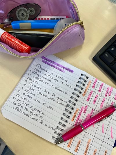 The image shows a notepad with handwritten notes in French, a pink pen resting on top, a pencil case, and part of a keyboard. The notepad lists steps or processes related to pharmaceuticals, including "units of care," "UFH service," and "selecting a criterion." It also contains highlighted date entries. The open pencil case reveals various writing tools, including a red lip balm, a white liquid highlighter, and a blue pen. A black keyboard is visible in the upper left corner.