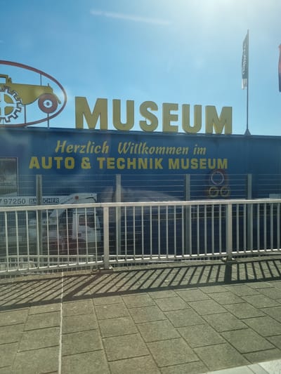 The image shows the entrance to the Auto & Technik Museum in Sinsheim, Germany. The building has a blue facade with the word "MUSEUM" written in large, gold letters. Below that, it reads "Herzlich Willkommen im AUTO & TECHNIK MUSEUM" (Welcome to the Auto & Technik Museum) in smaller gold letters. There's a graphic element featuring a stylized car shape and gears. A flag and banner are above the museum, visible against a bright blue sky.  A structure of vertical and horizontal metal bars creates shadows on the right side of the image and the facade of the building, with what appears to be a brick or stone wall further to the right. A small truck is visible through the metal bars behind the museum sign. An ad is also visible.